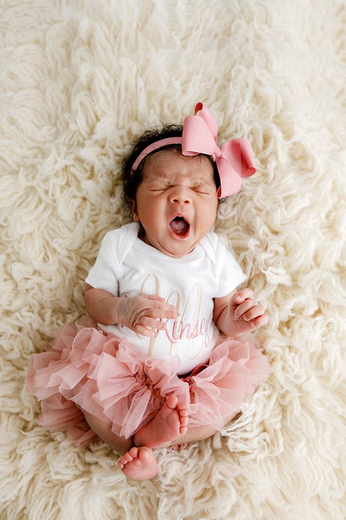 Yawning newborn baby girl in pink tutu and bow headband lying on fluffy white rug.