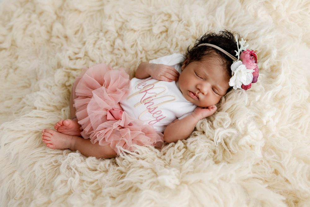 Sleeping newborn baby girl in pink tutu and floral headband on fluffy cream rug.