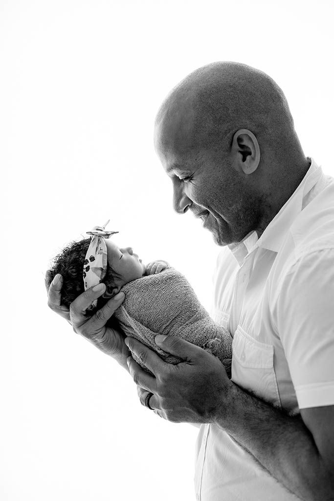 Black and white photo of a bald man smiling at his swaddled newborn baby by Perris newborn photographer.