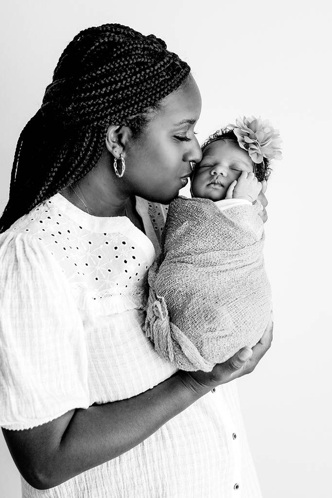 Black and white photo of a mother with braids kissing her swaddled newborn baby girl by Perris newborn photographer.