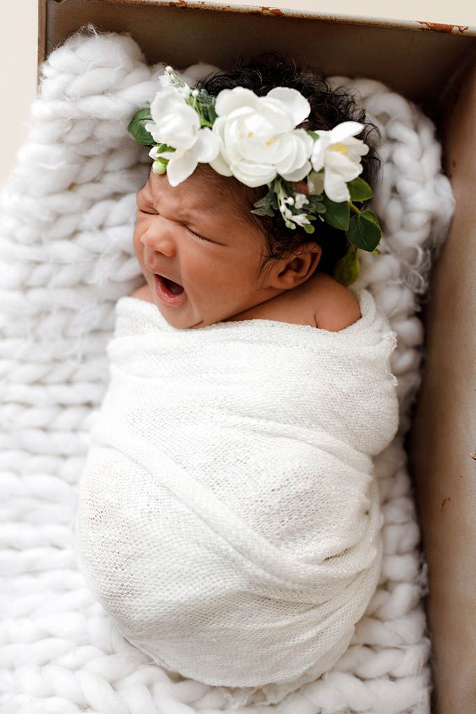 Newborn baby swaddled in white wrap, wearing floral crown, yawning peacefully in a cozy knit-lined box.