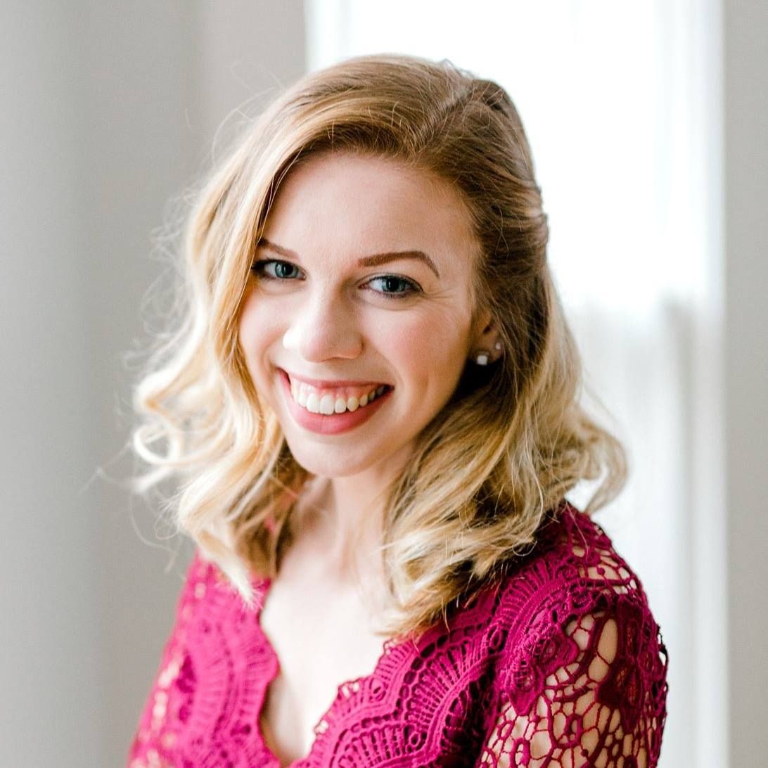 Smiling blonde woman in pink lace top poses for professional headshot photo near bright window.