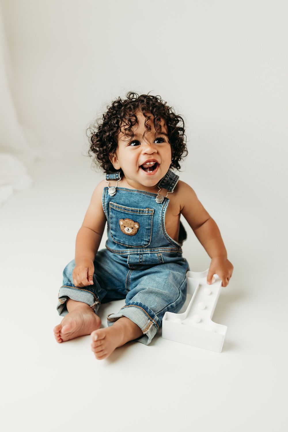 Smiling baby in denim overalls with bear patch sits barefoot on white background with number one prop.