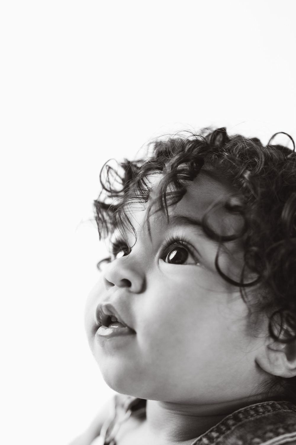 Black and white portrait of a curious toddler with curly hair looking upward against white background.