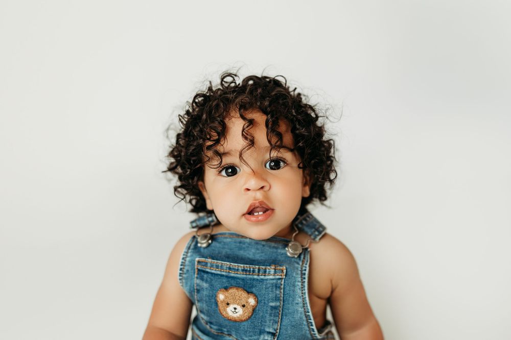 Adorable baby with curly hair wearing denim overalls with a bear patch, posing against a white background.