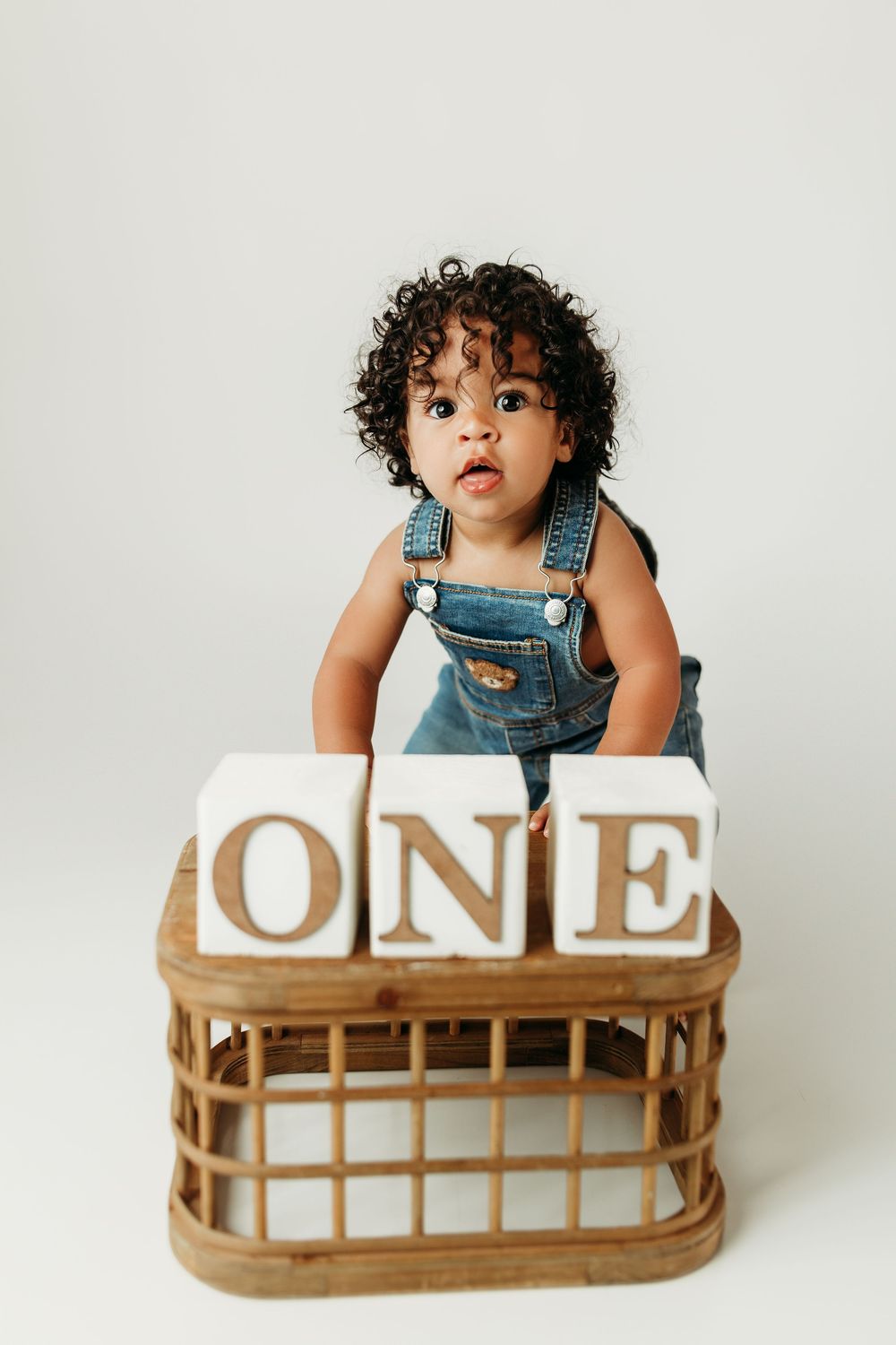 A toddler in denim overalls stands behind wooden ONE blocks in a wicker basket, celebrating their first birthday.