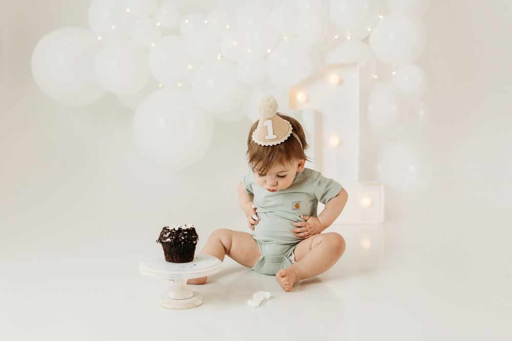 Baby in sage onesie sits beside chocolate smash cake with white balloon arch and number one backdrop for first birthday.
