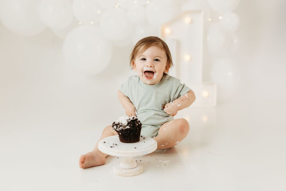 Baby's first birthday cake smash photo with chocolate cake, white balloons, and fairy lights on white background.