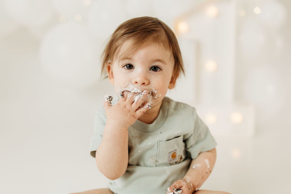 Baby boy in sage green shirt eating birthday cake smash with frosting on face and hands.