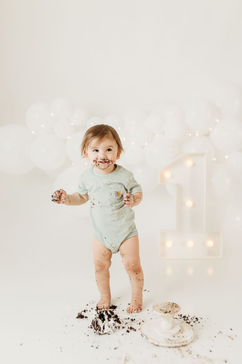 Smiling toddler in mint onesie standing in cake smash mess beside illuminated number one balloon backdrop.
