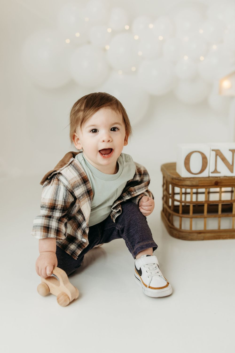Smiling baby boy in plaid shirt and Nike sneakers sits on white floor near wooden toy car and ONE sign.