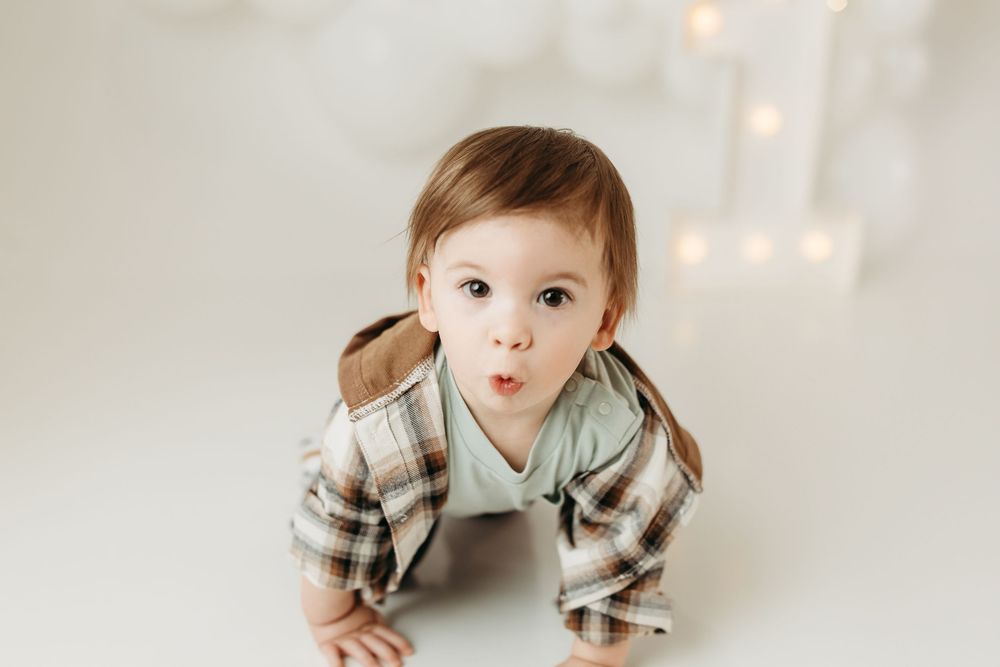 Adorable baby in plaid shirt crawling on white floor, looking up with surprised expression and bokeh lights background.