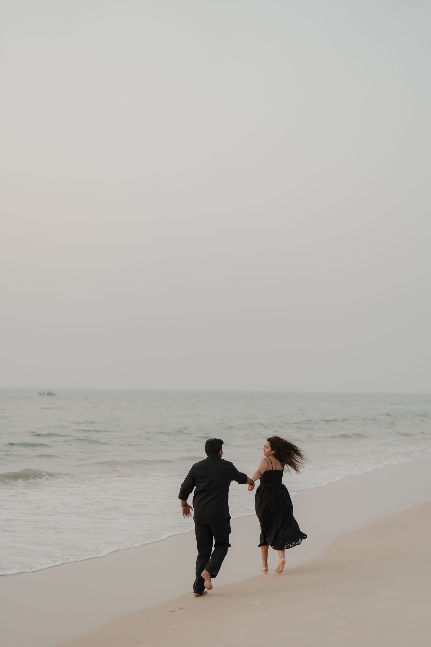 Black and white photo of two people holding hands on a sandy beach with gentle ocean waves in the background.