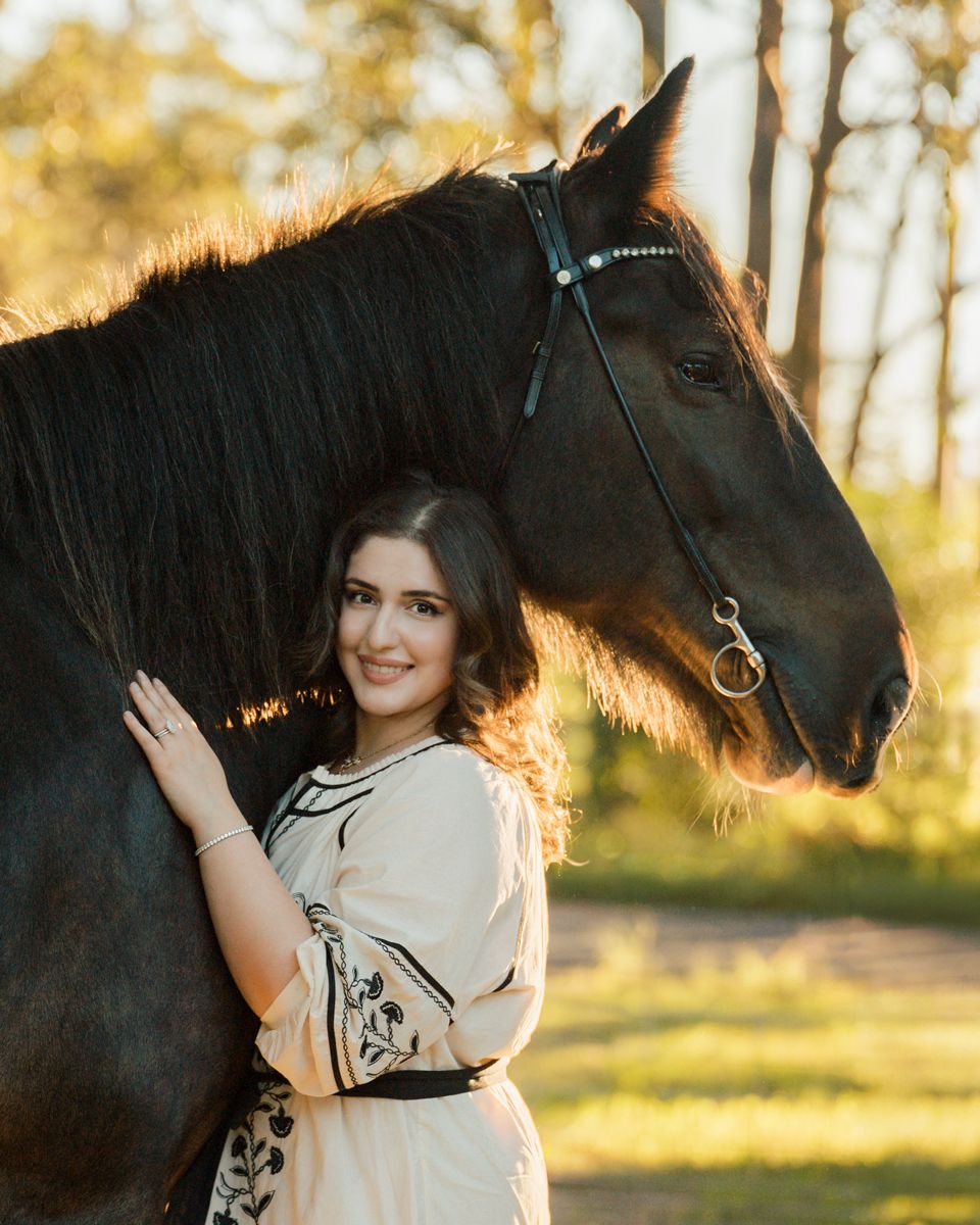 Young woman in floral dress smiling beside a black horse in a sunlit outdoor setting during an equine photoshoot in sydney hills district