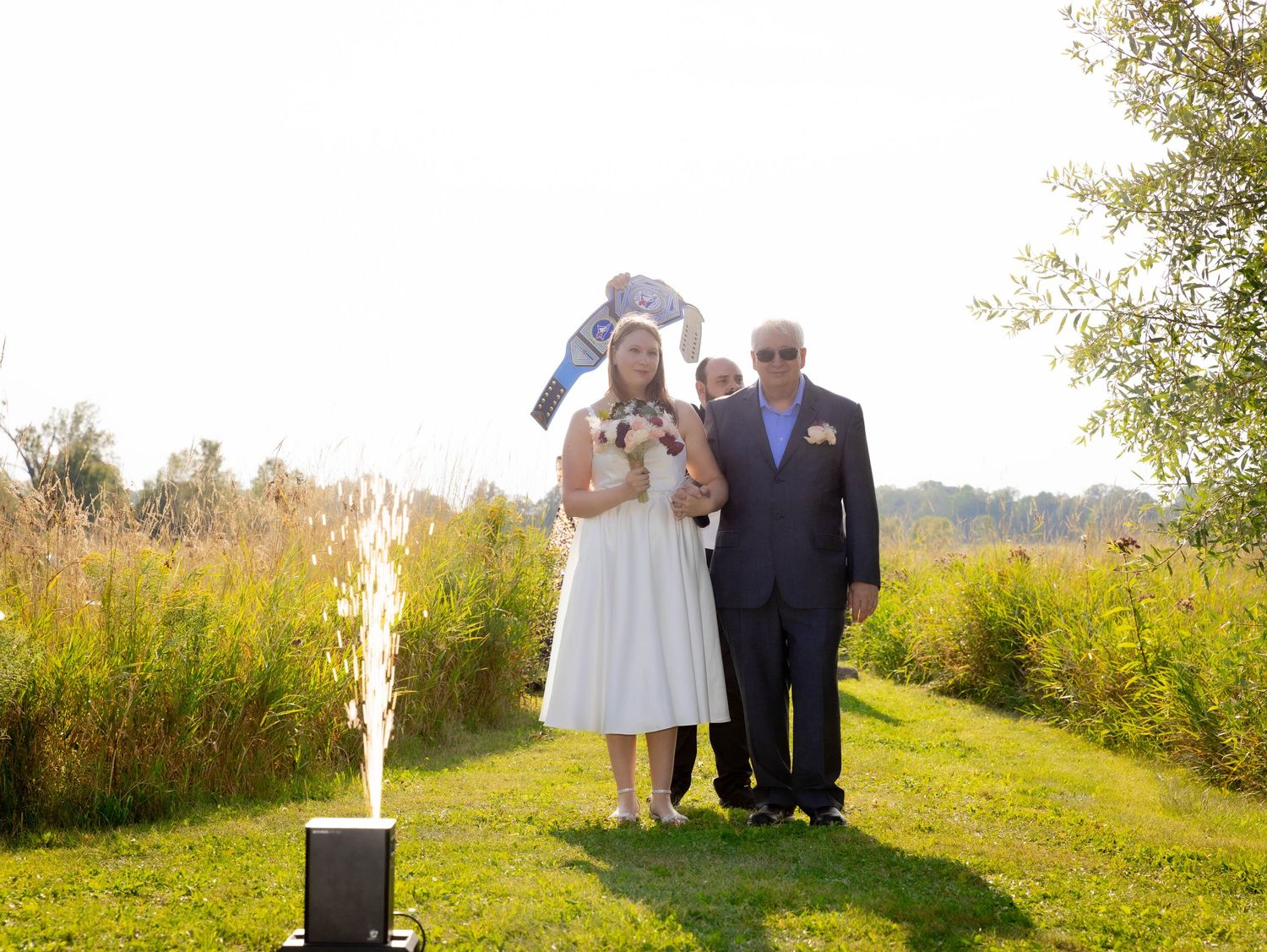 Woman in floral dress holds toddler wearing WWE championship belt outdoors, surrounded by bubbles near evergreen trees.