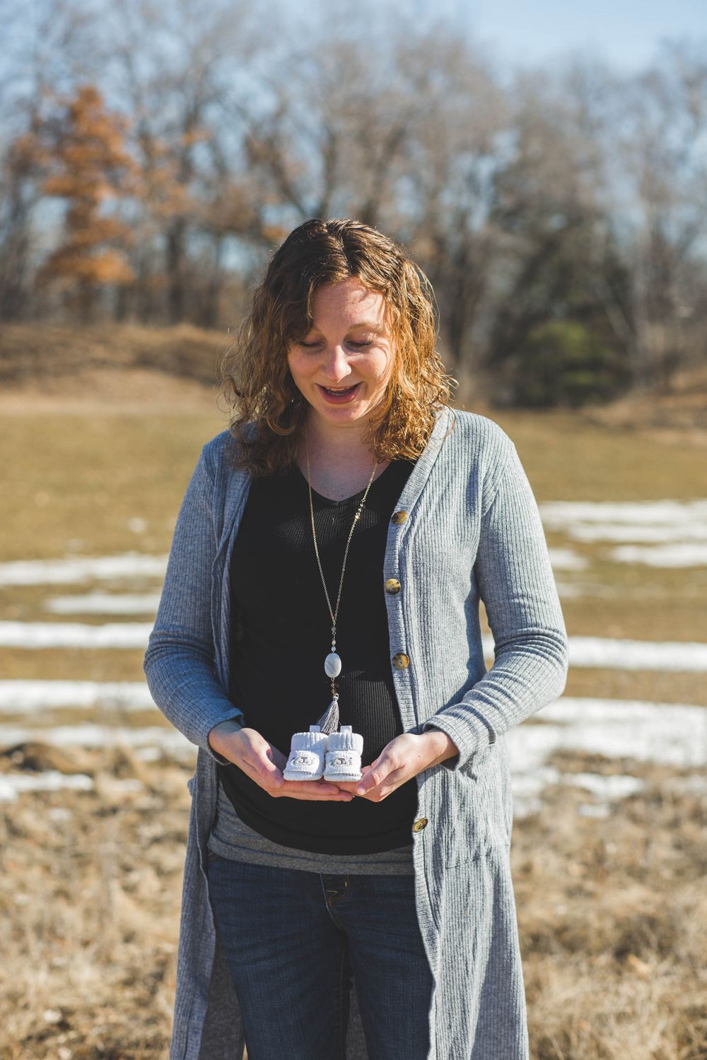 Smiling pregnant woman holding tiny baby shoes outdoors in a sunny field with patches of snow in early spring.