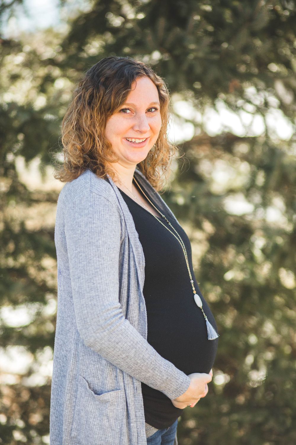 Smiling pregnant woman in gray cardigan and black top cradling baby bump outdoors near green trees.