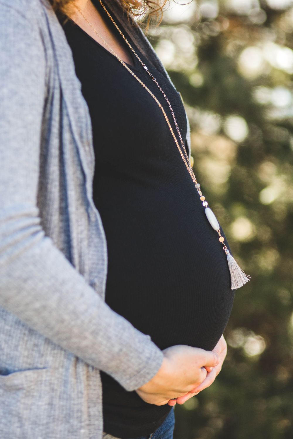 Pregnant woman in gray cardigan and black top cradling her baby bump outdoors, wearing a long beaded tassel necklace.