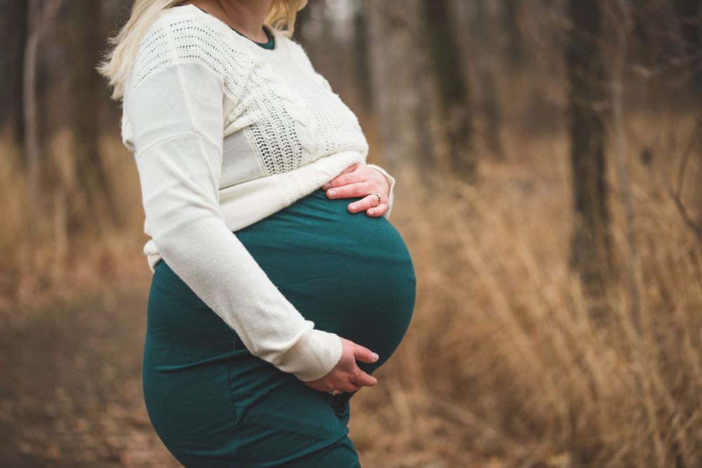 Pregnant woman in teal skirt and white sweater holding baby bump outdoors.