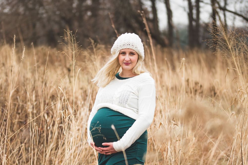 Pregnant woman in white sweater and teal skirt cradling bump outdoors.