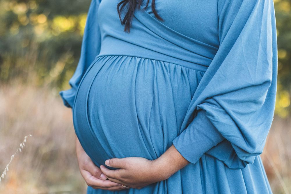 Pregnant woman in a flowing blue dress cradling her baby bump outdoors in a serene nature setting.