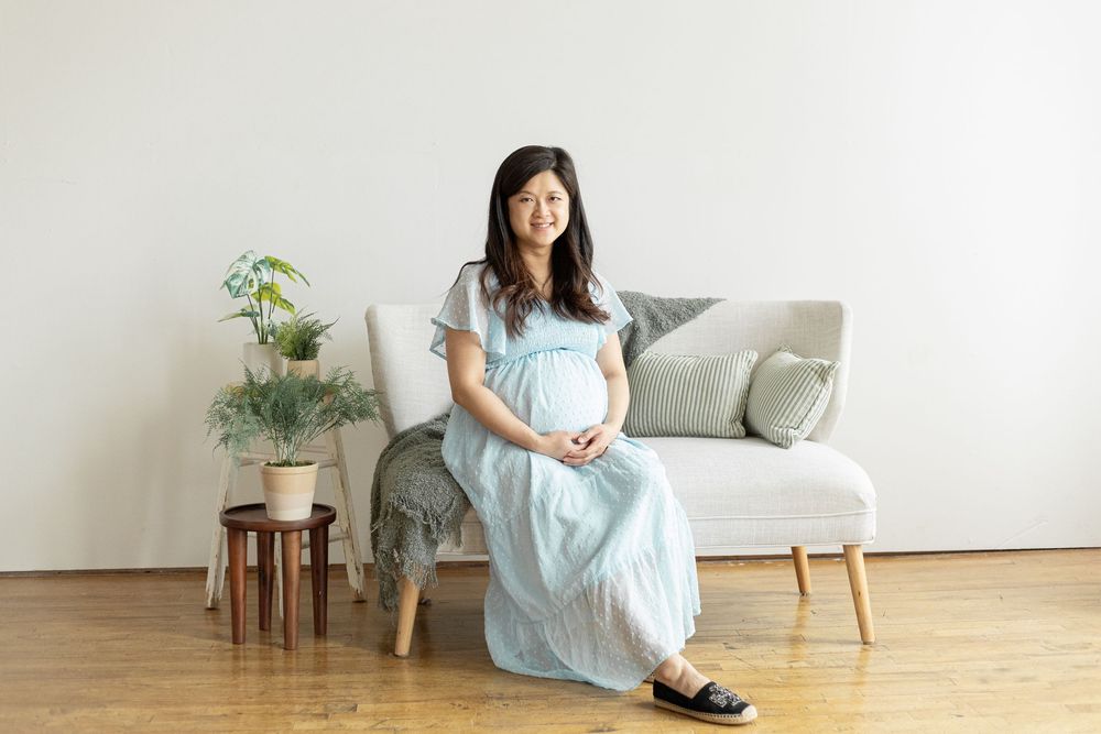 Smiling pregnant Asian woman in light blue maxi dress sitting on gray sofa in bright minimalist living room.