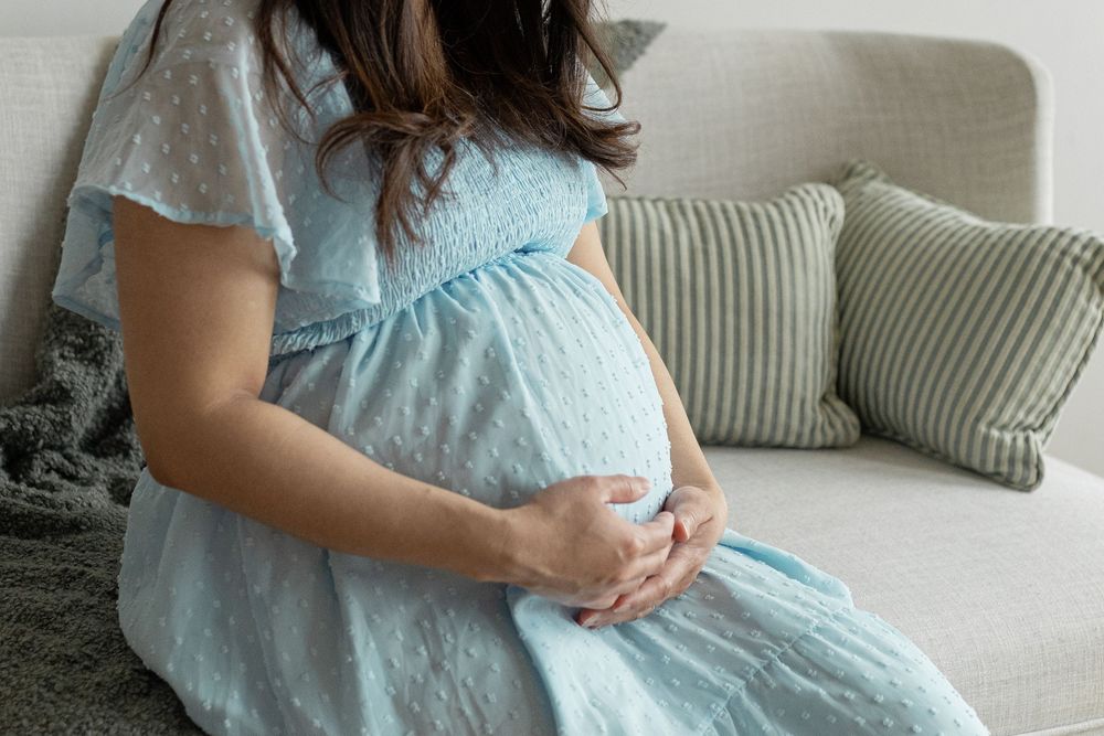 Pregnant woman in light blue dress sitting on sofa, cradling her baby bump with both hands.