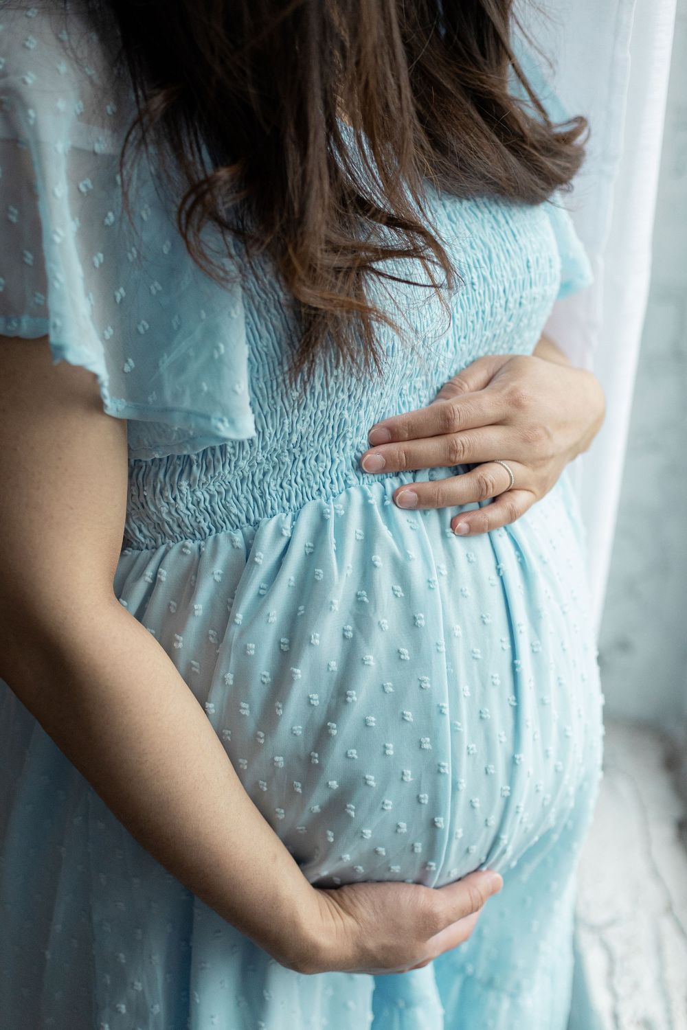 Close-up of pregnant woman in blue polka dot dress tenderly holding her baby bump with both hands.