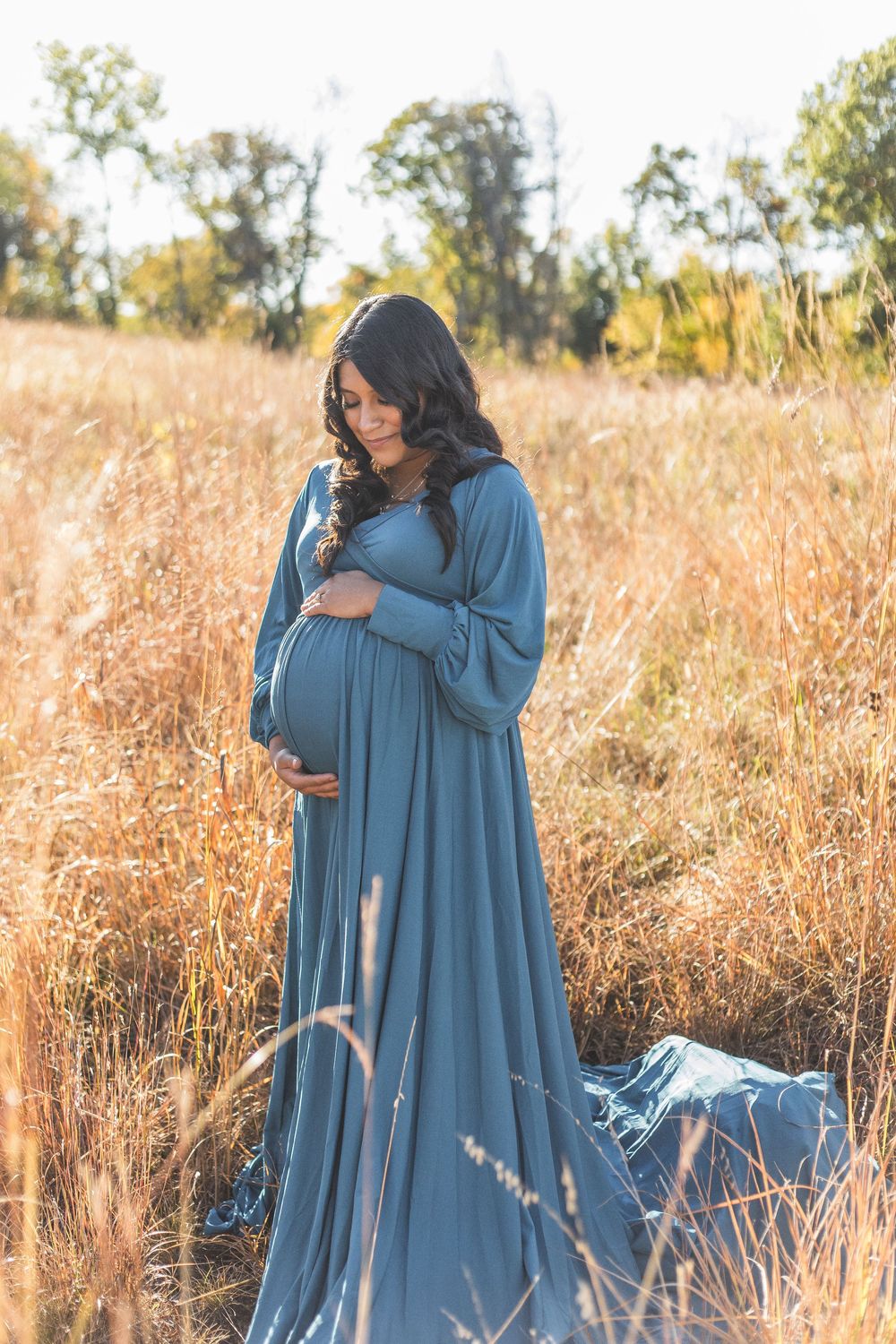 Pregnant woman in teal gown cradling baby bump in golden field during autumn maternity photoshoot.