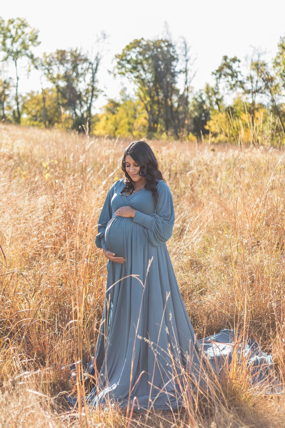 Pregnant woman in blue gown standing in golden field, cradling baby bump during outdoor maternity photo session.