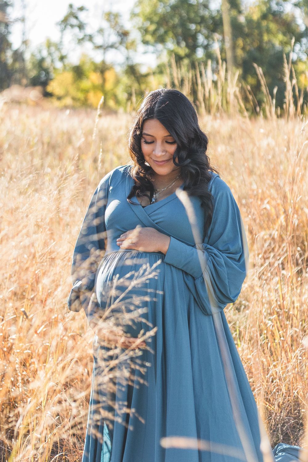 Pregnant woman in teal dress holding belly in golden field.