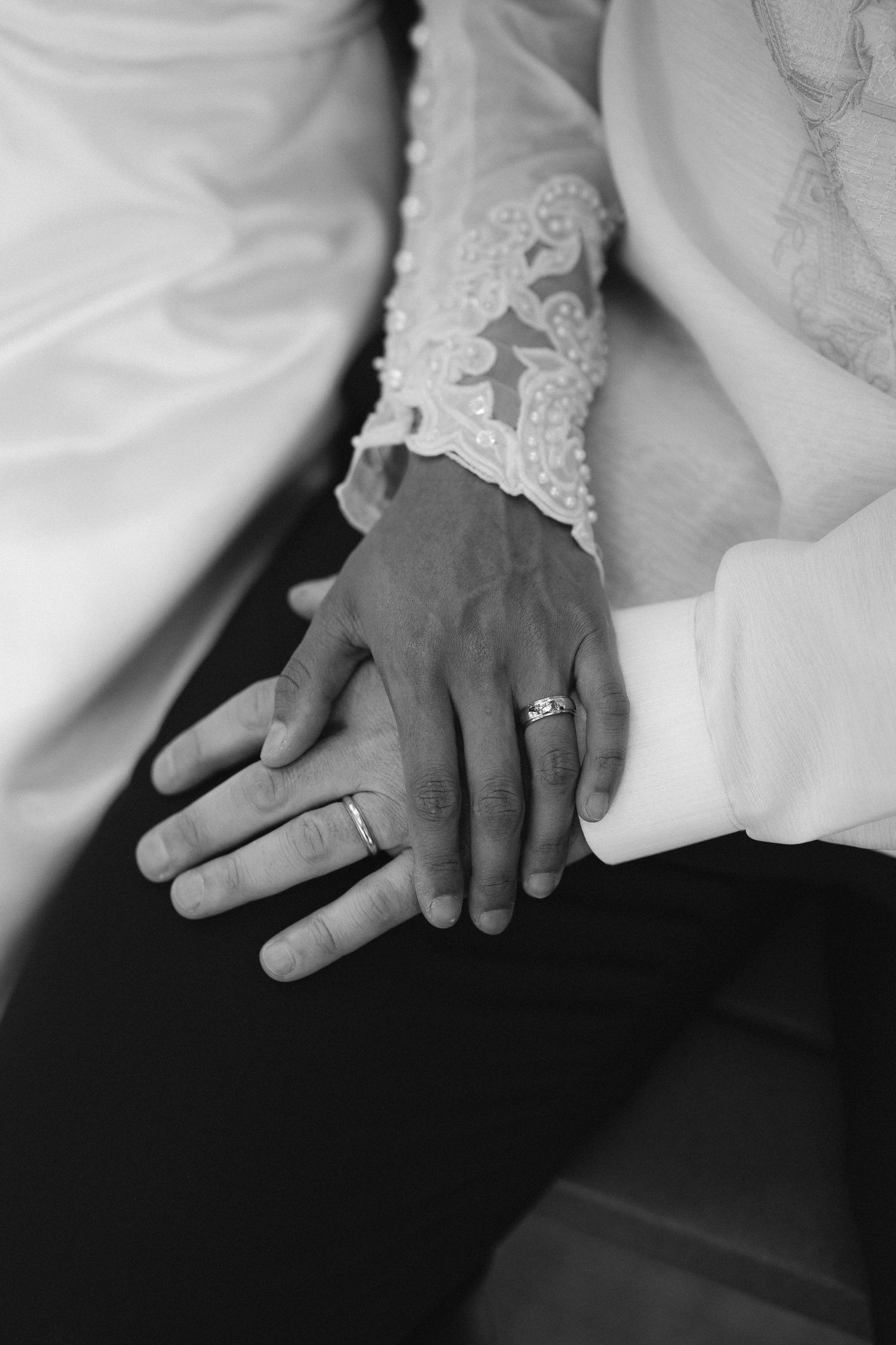 Black and white photo of newlyweds hands with wedding rings, bride in lace sleeve gown resting on grooms hand.