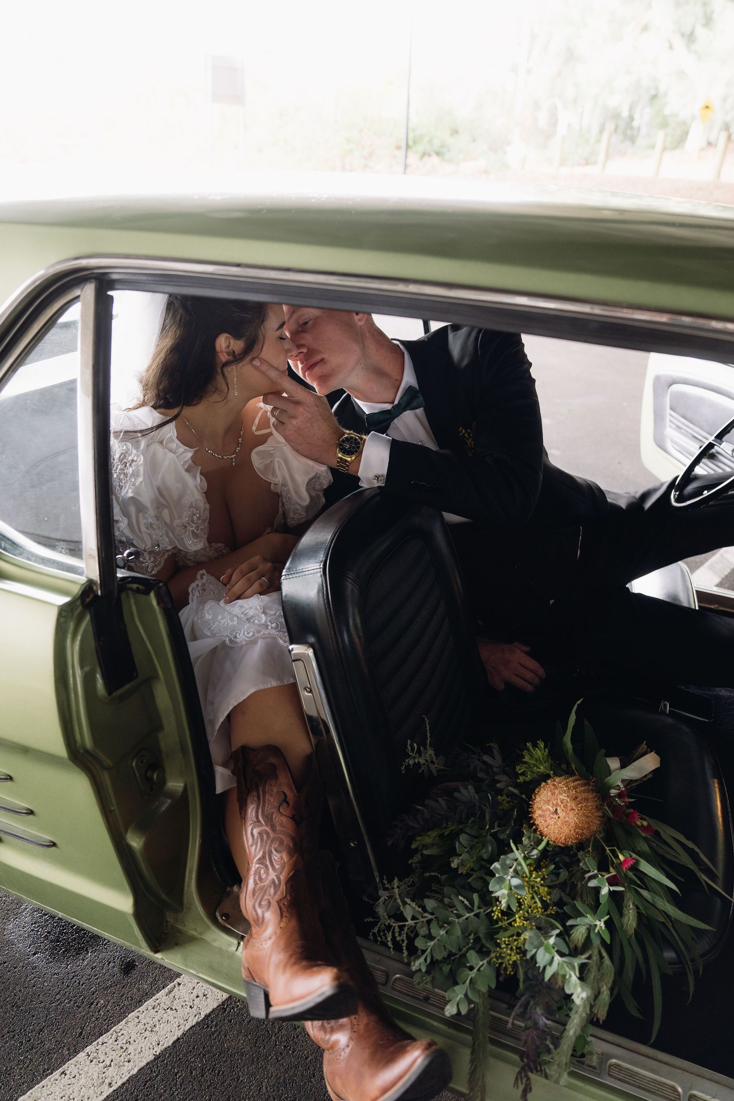 Bride and groom sharing a romantic moment in a vintage green car, with a floral bouquet and cowboy boots.