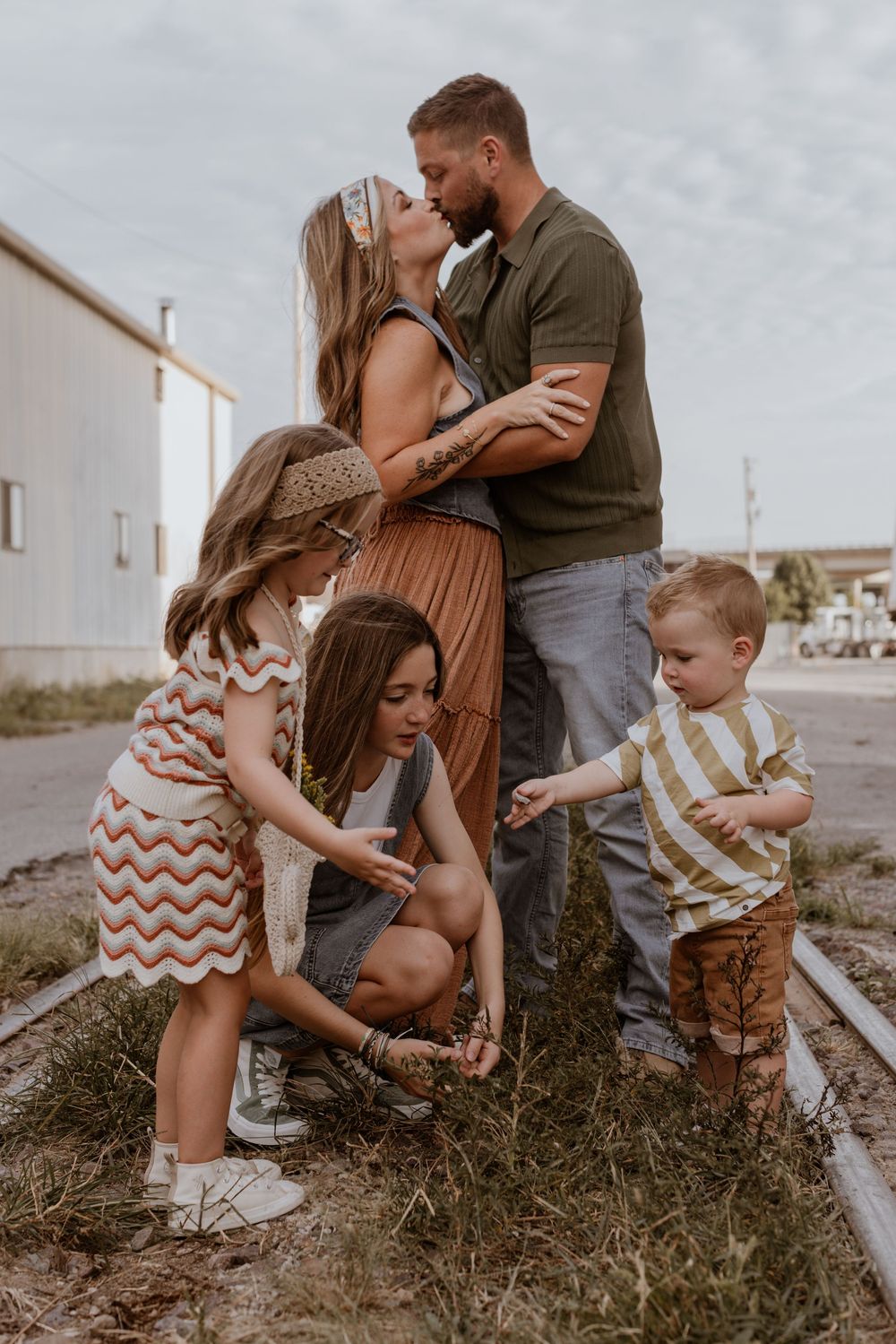 A mom and dad kiss in the background as their three children play in front of them during a Kansas City family photo session.