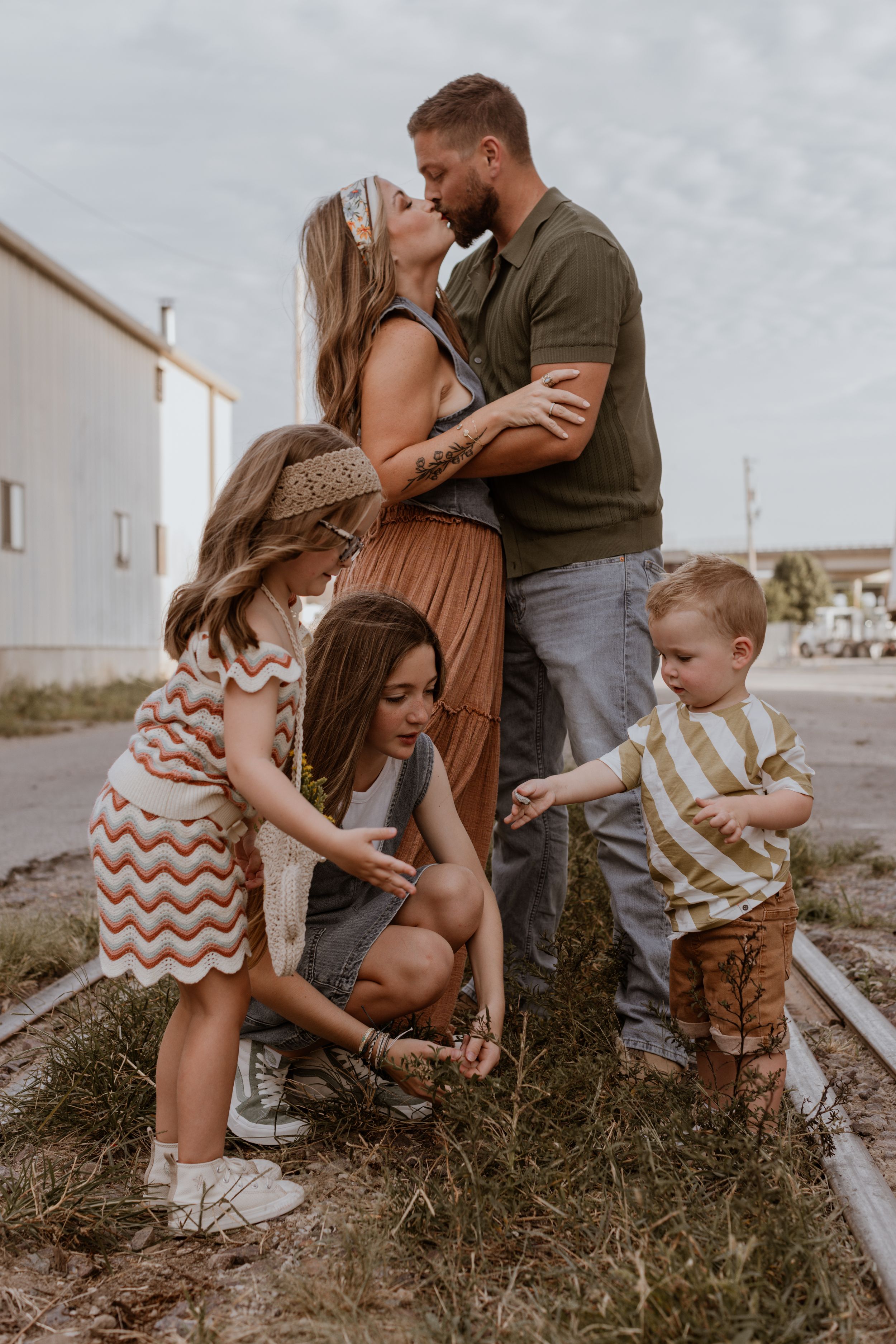 A mom and dad kiss in the background as their three children play in front of them during a Kansas City family photo session.