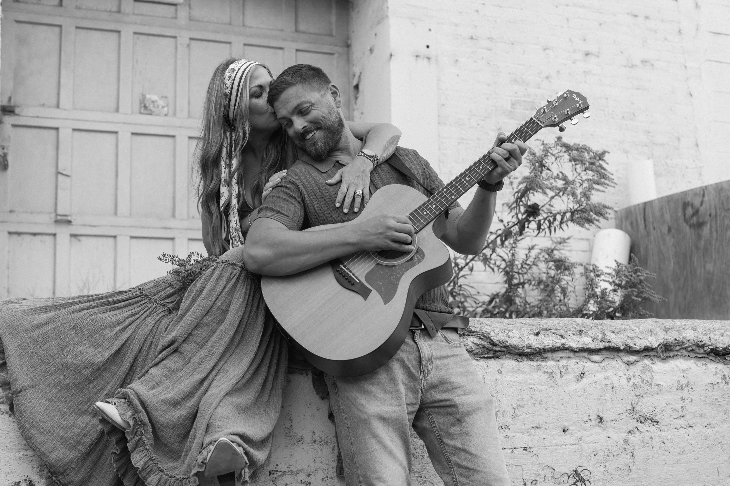 A father plays guitar and smiles as his wife lovingly embraces him from behind and kisses the side of his head during family photos.