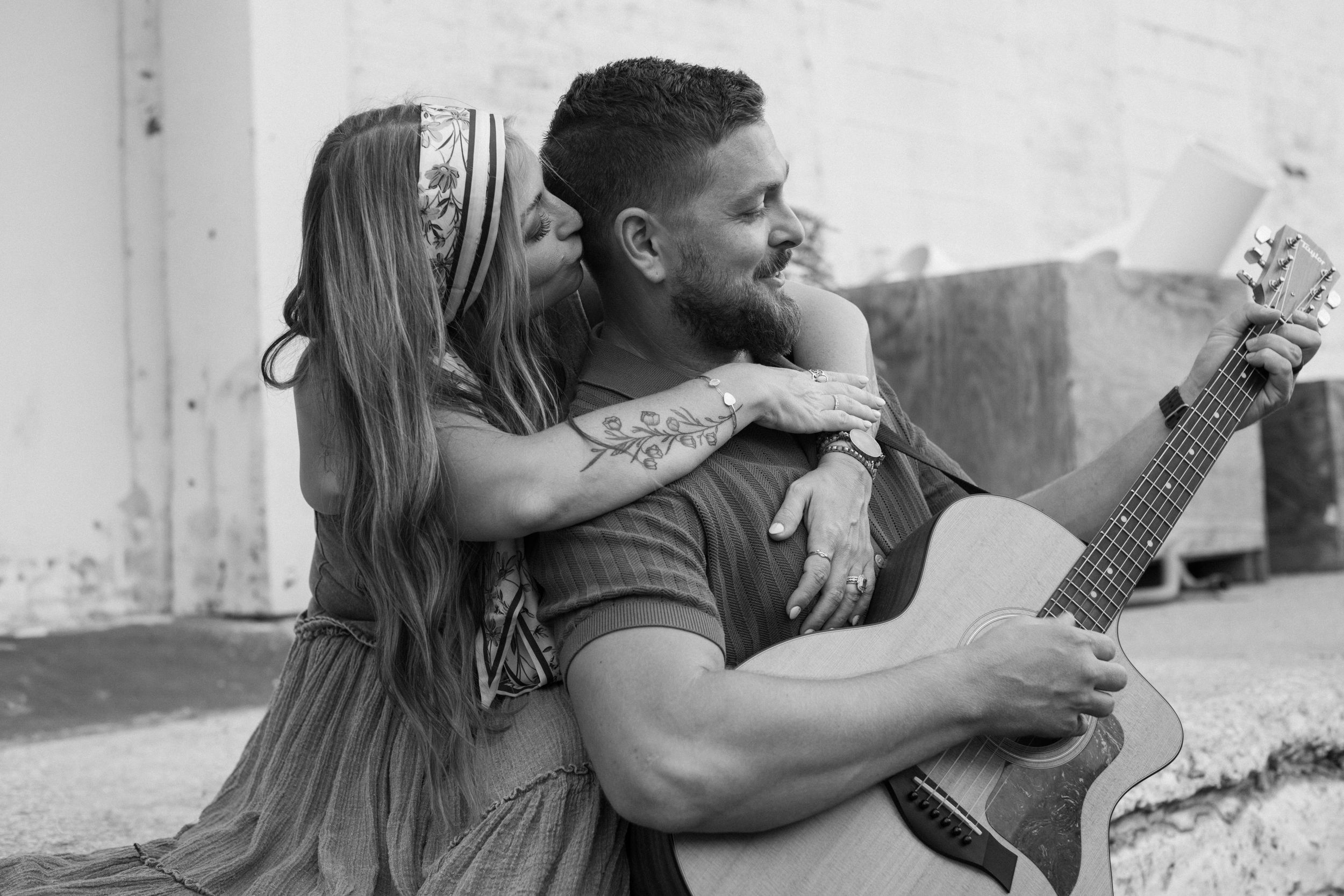 A father plays guitar and smiles as his wife lovingly embraces him from behind and kisses the side of his head during family photos.