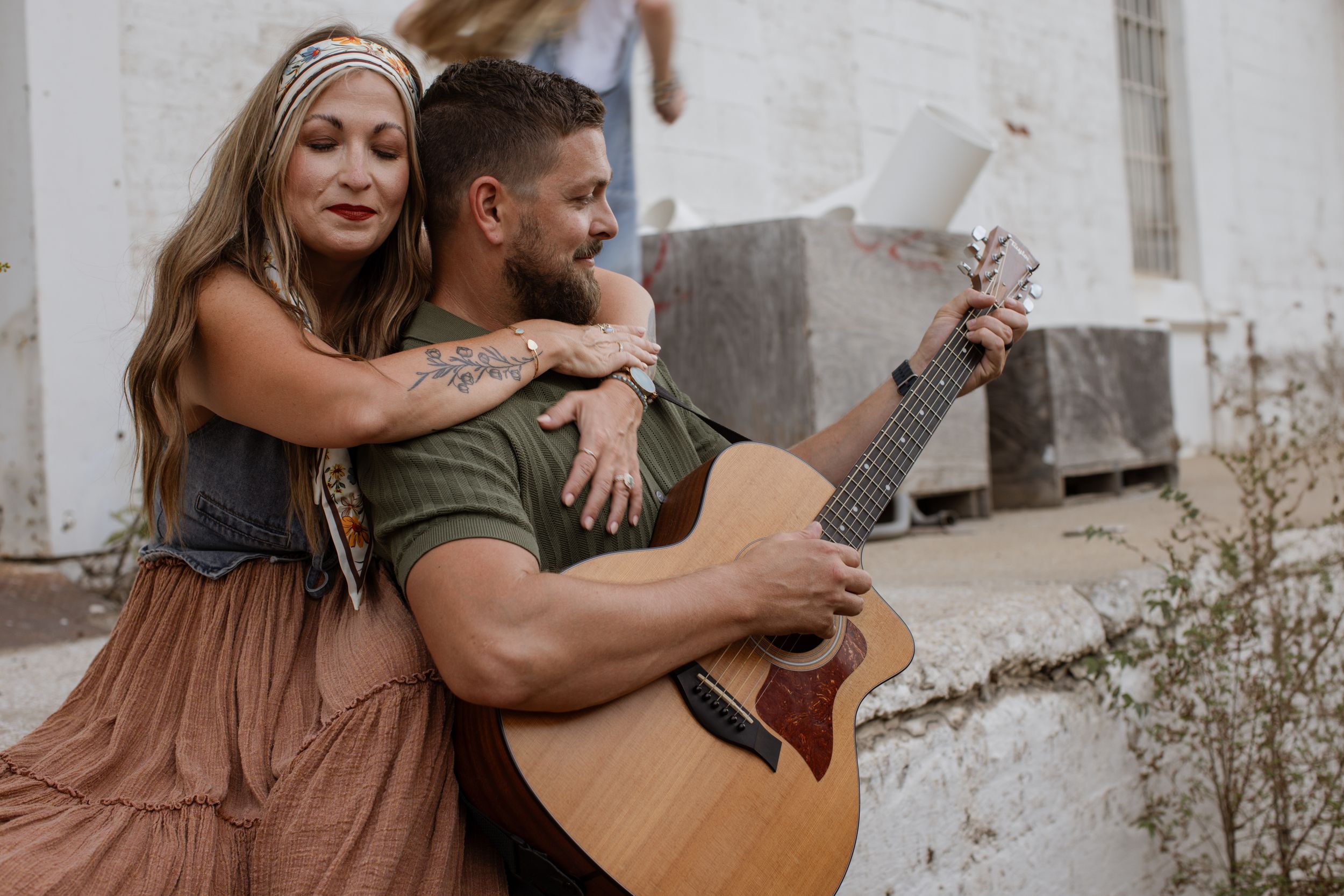 A father plays guitar and smiles as his wife lovingly embraces him from behind and closes her eyes during a Kansas City family photo session