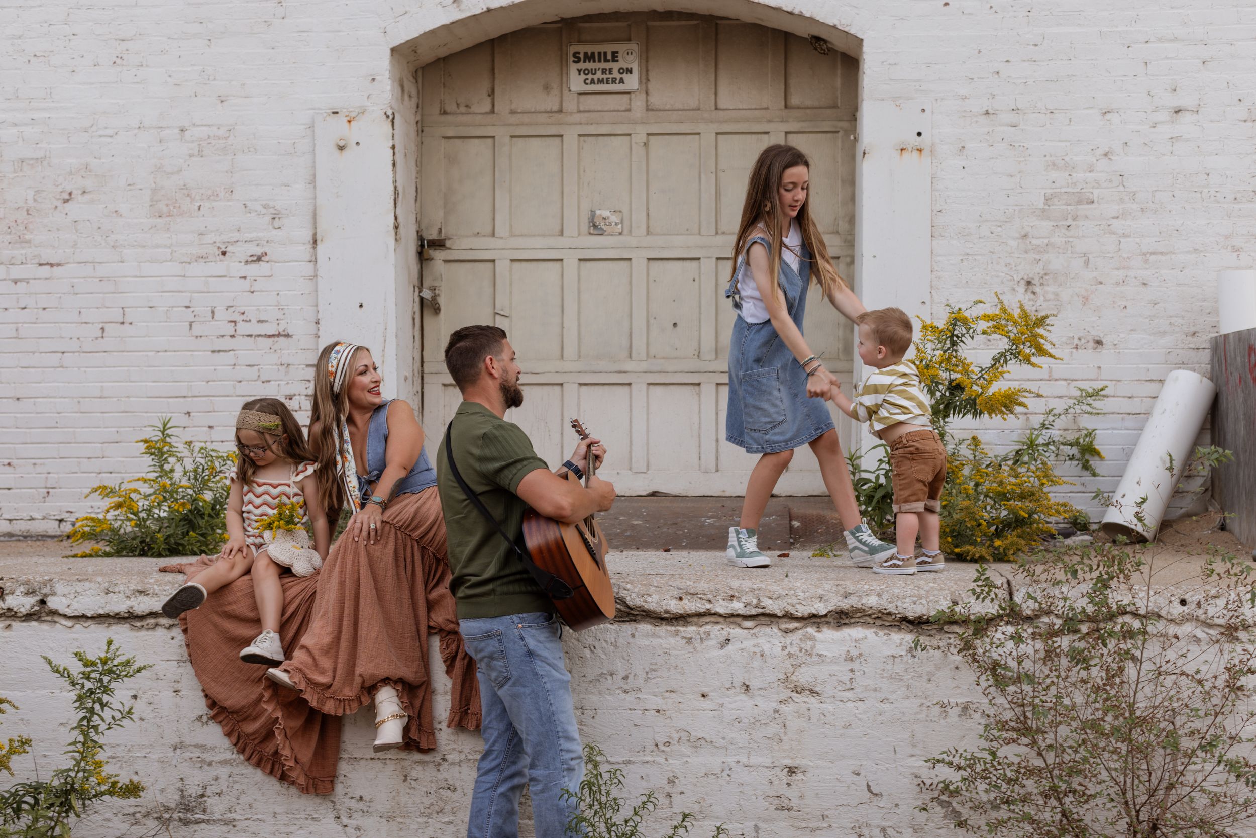 A father plays guitar while his teenage daughter and toddler son dance, their mother and sister looking on lovingly.