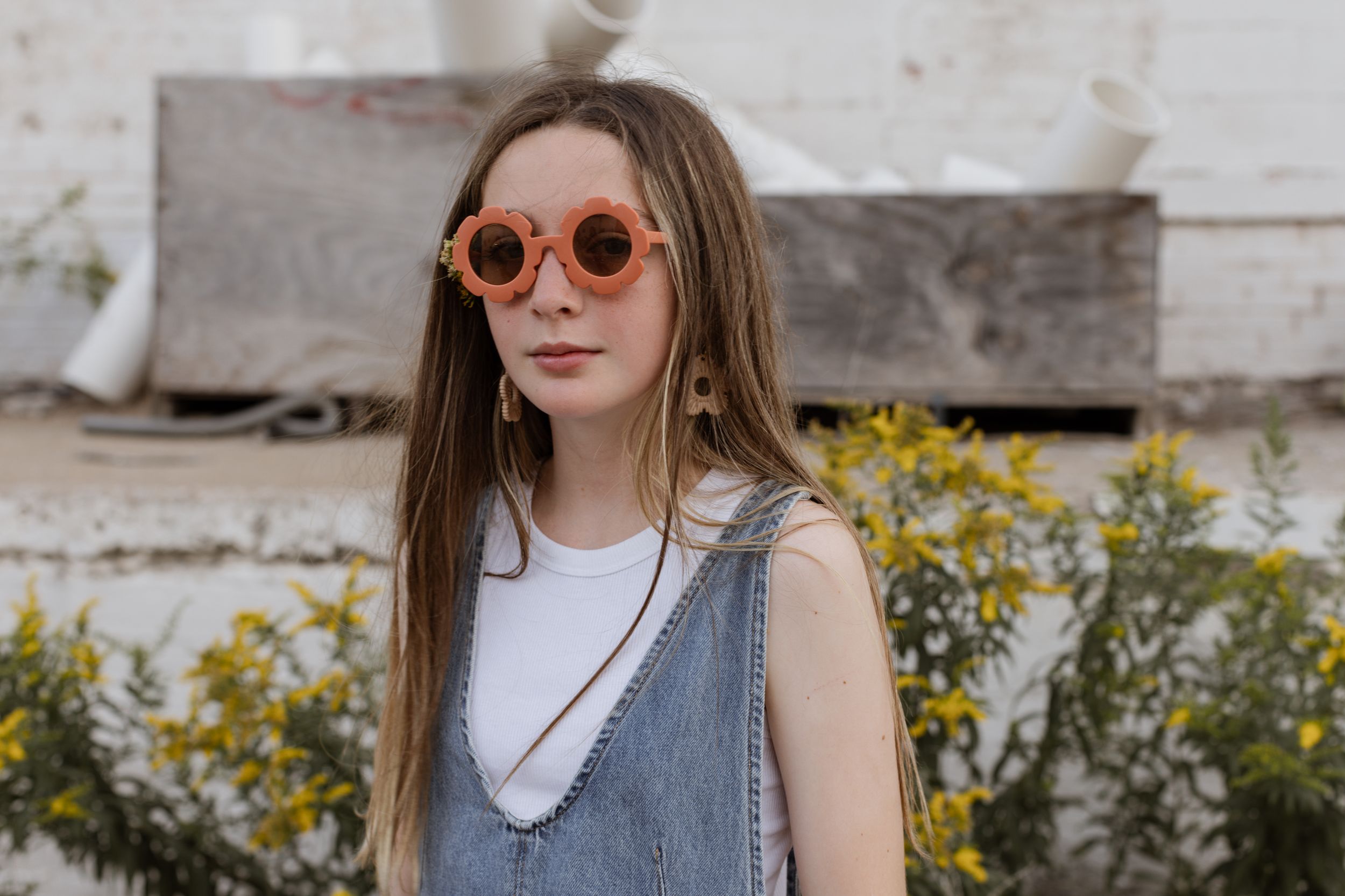 A young teenager wearing flower sunglasses looks at the camera with a white-washed building and yellow wildflowers behind her.