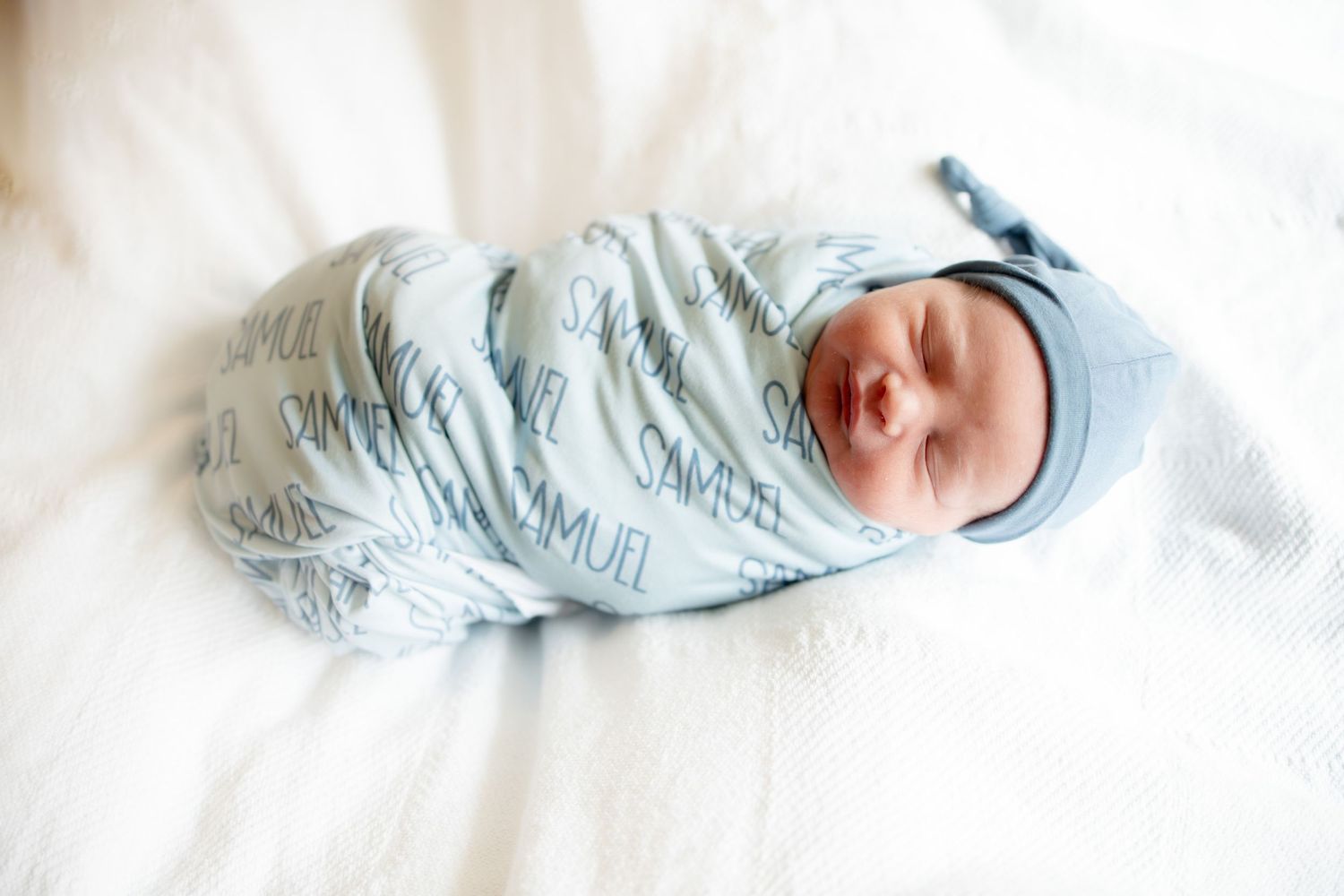 Newborn baby swaddled in personalized blue Samuel name print blanket with matching knotted hat.