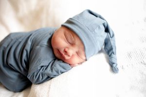 Sleeping newborn baby in blue footed pajamas and matching knotted hat on white blanket.