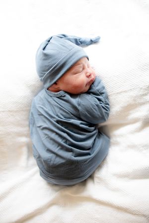 Newborn baby in blue sleeper and knotted cap sleeping on white blanket.