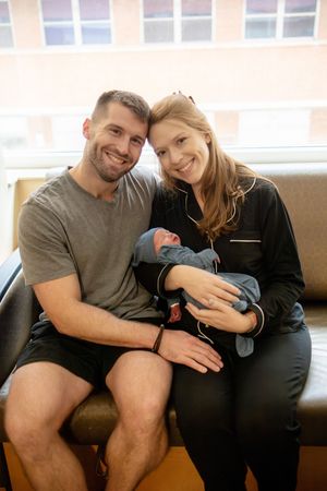 Smiling new parents posing together holding their newborn baby in hospital room.