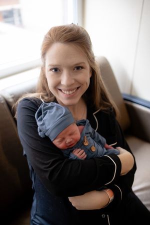 Smiling new mother holding newborn baby in blue outfit and hat in hospital room.