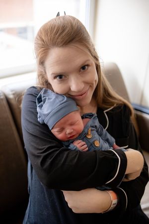New mother smiling while cradling sleeping newborn baby dressed in blue against her chest in hospital room.