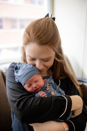 Young mother with red hair tenderly holding sleeping newborn baby in blue outfit and hat.