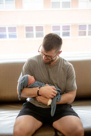 New father in gray shirt and glasses sitting on couch holding swaddled newborn baby in hospital.