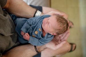 Close-up of sleeping newborn baby in blue outfit with wooden buttons cradled in parents hands.