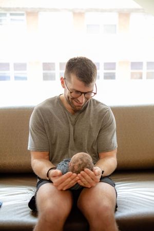 Emotional new father wearing glasses gently holding newborn babys head in his hands on couch.